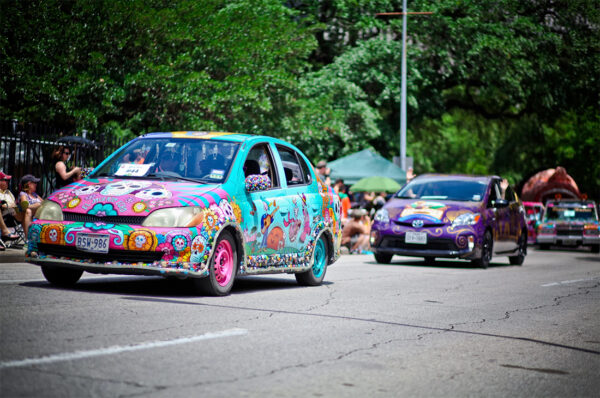 A brightly painted car participates in the Houston Car Art Parade. The four-door car is painted bright pink and turquoise and filled with Day of the Dead candy skull, calaveras, and marigold imagery. 