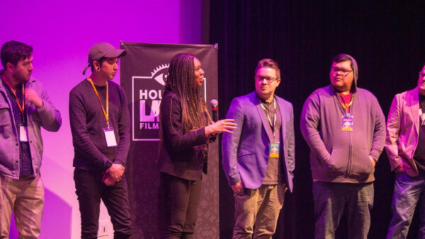 A photograph of a six people in conversation and standing on a stage. Behind them is a banner that reads, "Houston Latino Film Festival." The image is tinted purple due to the lighting.