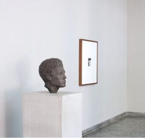 A slightly larger than life-size bust of the artist Alberto Giacometti sits on a white pedestal. To the right of the sculpture is a framed photograph. Installation by Teresa Hubbard and Alexander Birchler.