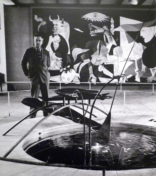 A black and white photo of Alexander Calder between a fountain and Picasso's painting of Guernica