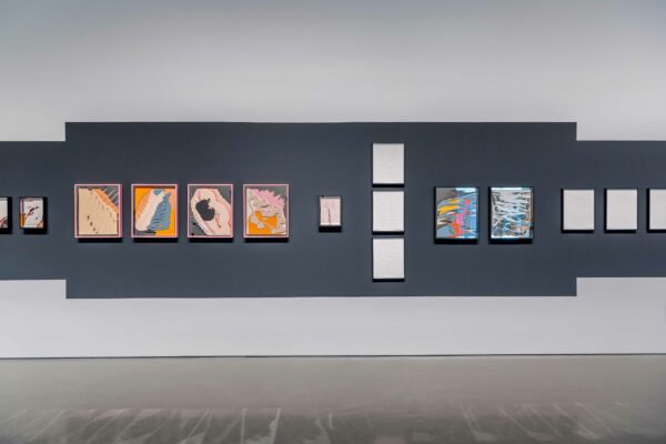 Image of multiple, colorful, framed works on paper against a gray wall.