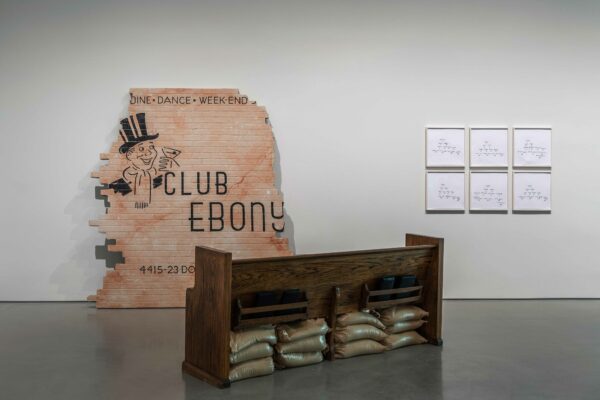 Installation view of a work by Jamal Cyrus. A partial, false brick wall leans agains the gallery wall in the background, and a short wood shelf with bags of dirt stand in front.