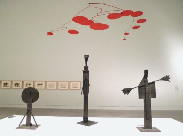 Exhibition view with work by Calder and Picasso. In the foreground are three bronze sculptures of figures, works on paper in the background, and a red mobile by Calder suspended from the ceiling.