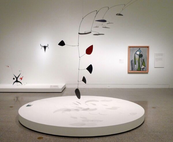 View of exhibition with works by Calder and Picasso. A red and black mobile by Calder hangs in the foreground and smaller sculptures and paintings by both artists are in the background