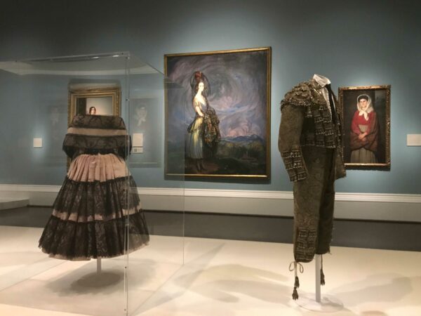 An installation photograph of a museum exhibition. The image features paintings on the museum's walls, and mannequin forms wearing clothes standing in the middle of the room