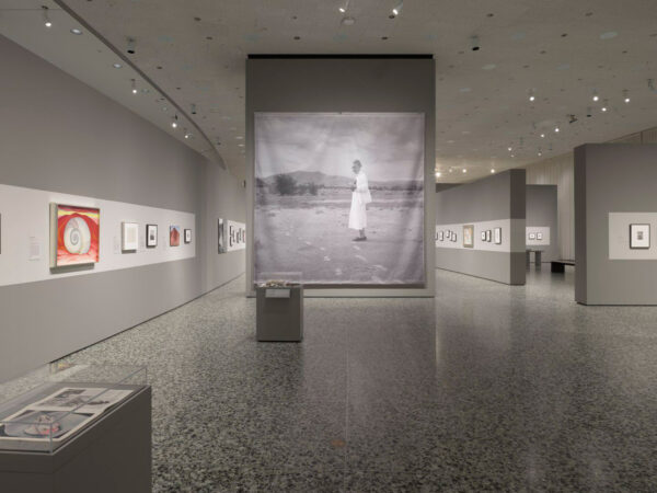 Photograph of an art show featuring photographs by artist Georgia O'Keefe. The room features a gray floor and walls, along with many, smaller framed artworks.