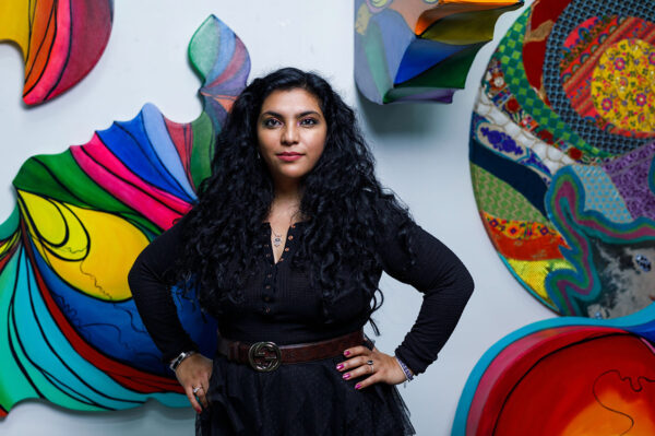 A photograph of artist Janavi Mahimtura Folmsbee standing in front of her large bright colorful paintings. The artist wears a black dress and stands with her hands on her hips. Her dark dress and black hair contrast against the vivid colors of the irregularly shaped paintings behind her.
