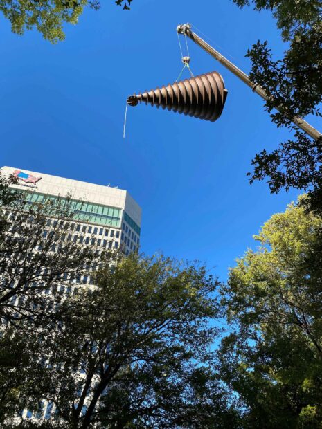 A crane hoisting a steel, conical-shaped steel sculpture up into the air.