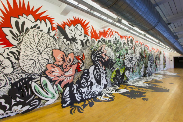 Installation view of a mural by artist Natasha Bowdoin that shows a layered and mnulti colored, large scale installation of flora agains a white wall.