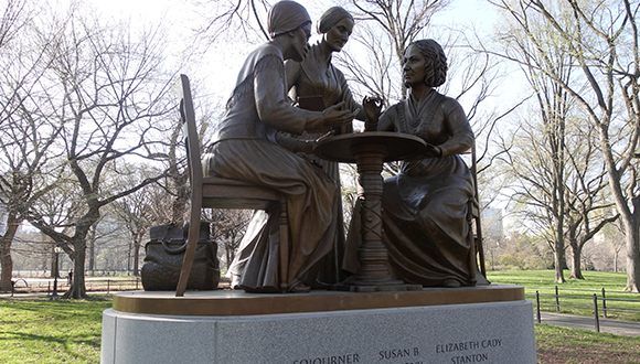 An outdoor, public monument surrounded by trees of three women at a table. The monument is for the pioneers of the women's movement