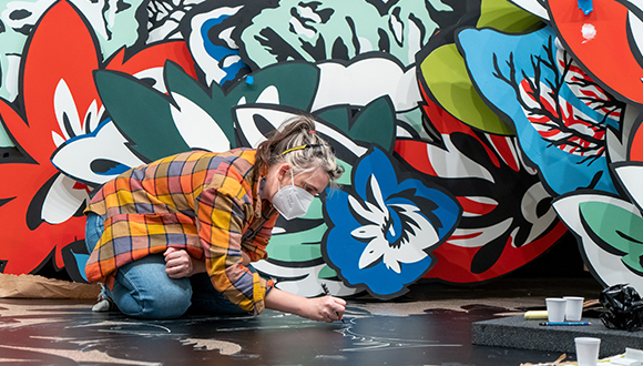 A photo of the artist working on a mural. The artist is kneeling and painting directly on the floor, parts of the large scale mural are shown in the background behind her.