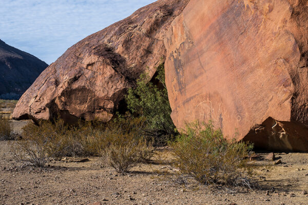 A color photograph depicting large red boulders. The boulder on the right has lightly etched petroglyphs. In front of the boulders the desert ground has sparse vegetation. Photo by Paul Leicht.