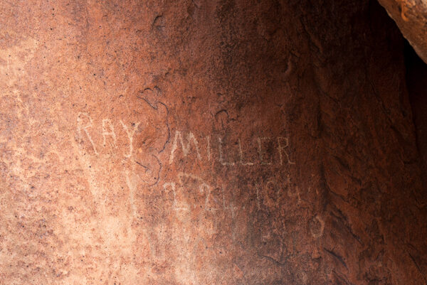 A close-up photograph of a red stone. The text, "Ray Miller 2.24.104" is etched on top of a petroglyph. 