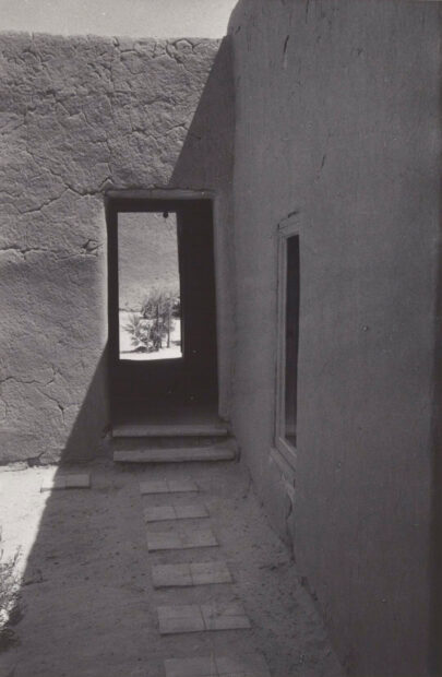 A photograph of a doorway of an adobe-looking house. The foreground features steps embedded in the ground, and through the doorway we see out another doorway onto trees.