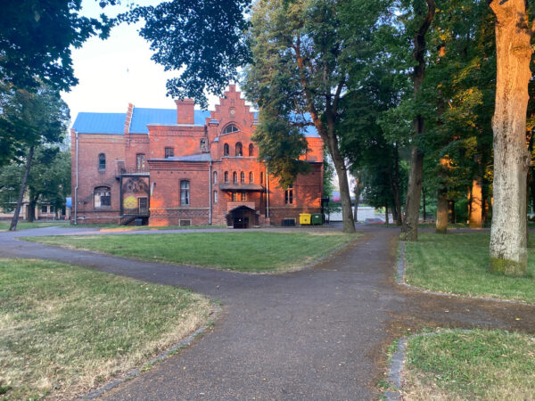 A photograph looking up the driveway of a large, house-looking building. The house is made of red brick and features many architectural adornments.