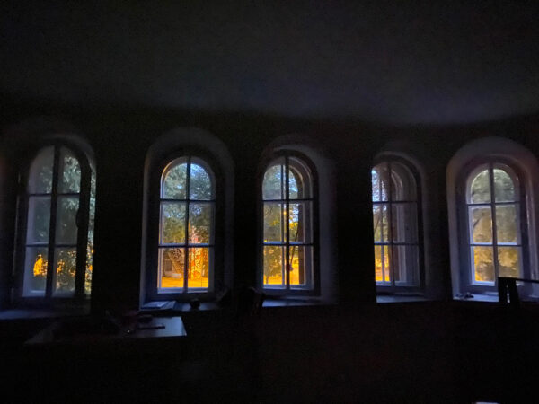 A photograph of the interior of a building. Through the building's five windows, you can see outdoors.