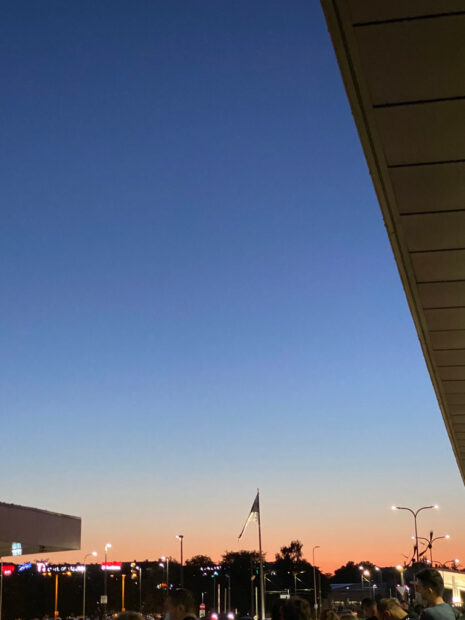 Photograph of a sky. The sky has a gradation of color and is primarily blue. There is a flagpole in the foreground, with an indistinct flag hanging from it.