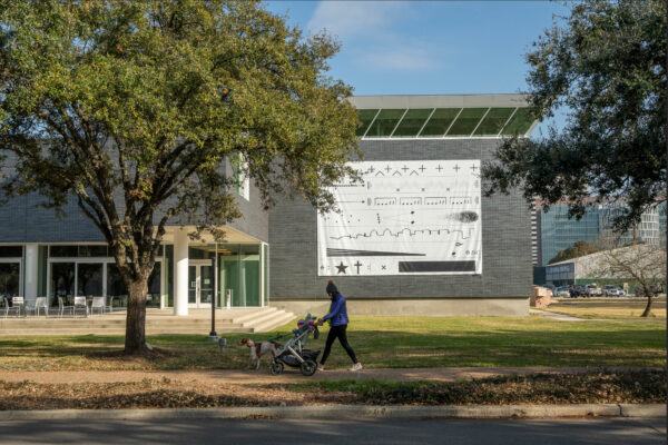 View of an outdoor piece hanging on the side of the Moody Center for the Arts at Rice University. The work is a tarp with various sound shapes printed on it.