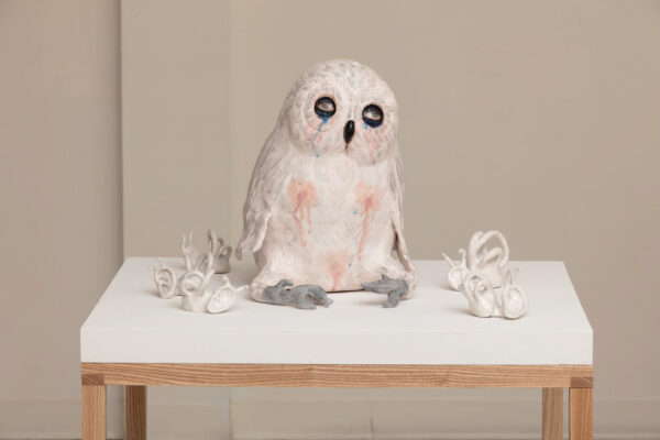 A photograph of a ceramic art installation. On a white table top, an owl sits surrounded by five small ceramic pieces that resemble a combination of talons and human ears. The owl's feathers are white, its talons are gray, and its beak and the areas surrounding its eyes are black. The eyes have been painted to look like human eyes and blue paint resembling tears drip down its face. Its chest and lower abdomen have spots of pink paint that also drip down its torso. Artwork by Margaret Meehan. 