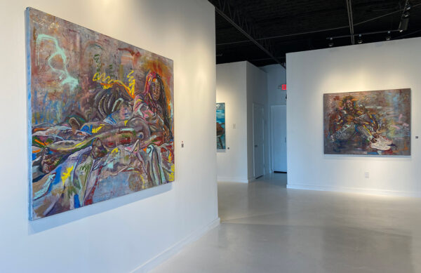 A photograph depicting the interior of a gallery. Two large horizontally oriented paintings hang on separate walls. Each painting uses expressive brushstrokes and bright colors to depict the subjects. The painting on the left is one a woman lounging and the painting on the right is of a man sitting. Artwork by Sharidyn Barnes.