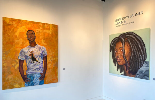 A photograph showing the entry wall of a gallery. Two large oil paintings are hung on adjacent walls. On the left is a painting of a male figure set against a bright orange background. On the right is a self-portrait with wall text that reads, "Sharidyn Barnes "Unseen," January 5 - February 5, 2022"