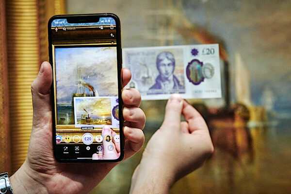 A photo of a museum visitor taking a photo of a 20 pound bill in front of a painting by JMW Turner