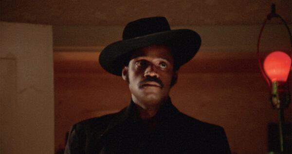 A film still with Melvin van Peebles facing the camera wearing a black cowboy hat tilted to his right side and a black trench coat.