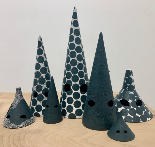 Photos of a group of 7 statues of different sizes.  Each component is cone-shaped with a pair of circles or almond-shaped holes resembling eyes.  Some works are painted completely black, while others are white and covered with black circles that are placed close together.  Drawing by Tammie Rubin.