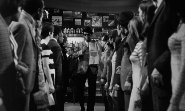 A film still of two rows of people looking at one man standing in the back and center between them.