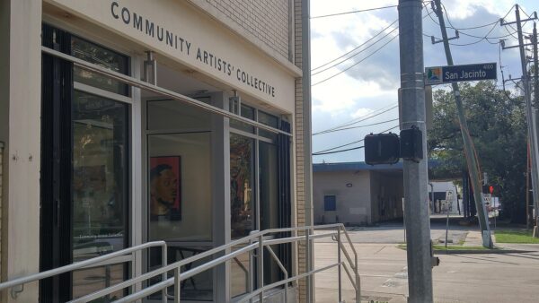 A photograph of a street corner. On the right you can see the facade of light colored brick building. Above the entrance, the sign reads, "Community Artists' Collective."