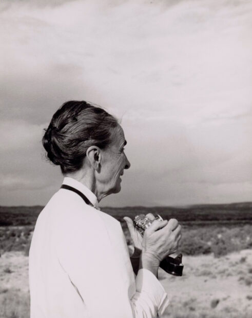 A photograph of artist Georgia O'Keefe. The artist is holding a camera in her hands, and looking out across a brush landscape.