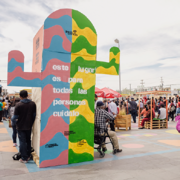 A photograph of a large cactus sculpture. The cactus is painted in unrealistic colors with pink and blue on the left and green and yellow on the right. White text is stenciled on top of the painted colors and reads, "este lugar es para todas las personas cuídalo." Artwork by Nómada Lab.