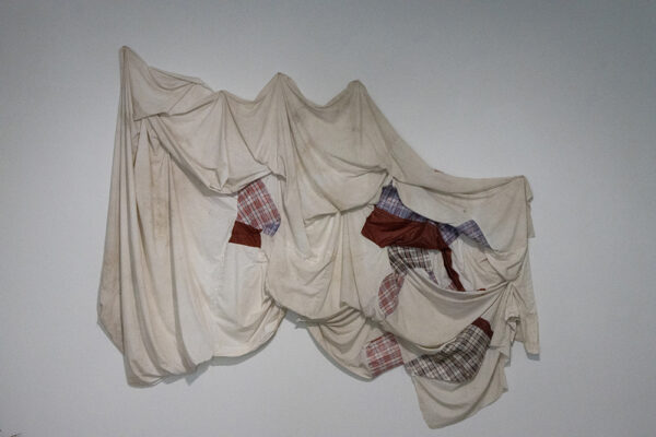 A sculptural piece hanging on a gallery wall. The work of art is created from mostly white fabrics but also includes some small patches of plaid. It resembles bedsheets and is pinned to the wall at various points along the top. Artwork by Verónica Gaona.