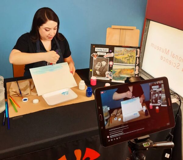 A color photograph of a woman sitting at a desk and demonstrating painting techniques. To her left is a board with a photograph of and paintings by artist Emily Carr. In the foreground there is a tablet creating a video recording of the instructor.