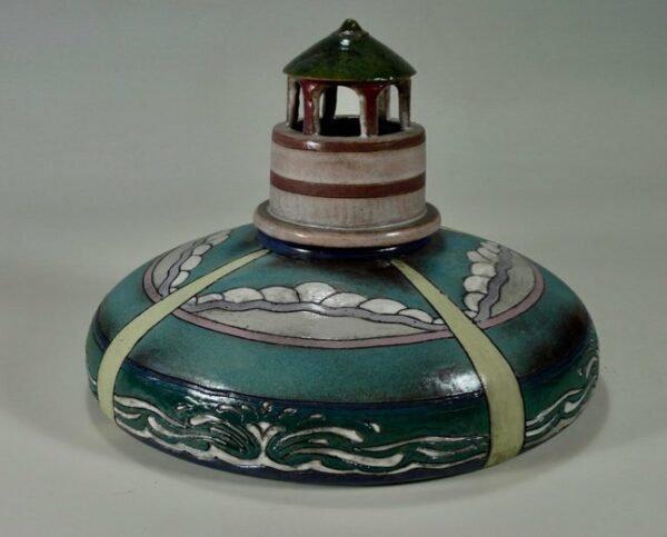 A photograph of a ceramic sculpture by William Tersteeg. The sculpture has a short, circular base with a short lighthouse standing in the middle. The edges of the base have depictions of the ocean. The top of the base resembles a rocky shore. 