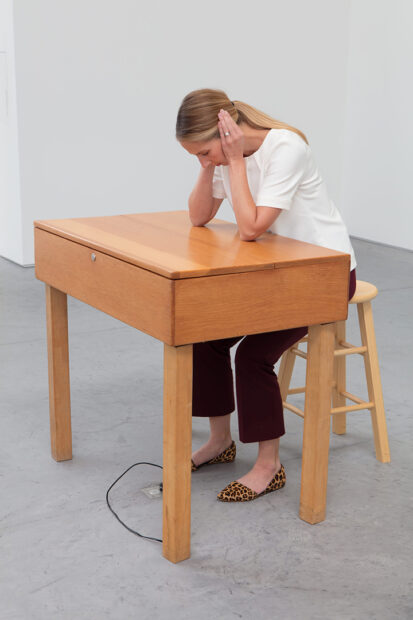 A photograph of a visitor interacting with a sound art work by Laurie Anderson. A woman sits on a wooden stool in front of a wooden table. Her elbows are on the table and her hands are covering her ears.