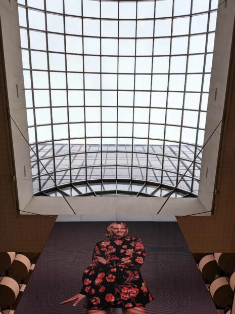 Photograph looking up, at a large skylight. In the foreground is a large screen, depicting a person wearing a floral outfit.