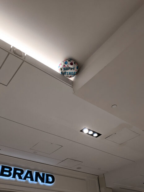 A photograph of a mylar balloon stuck at the top of a corner of the ceiling of a mall.