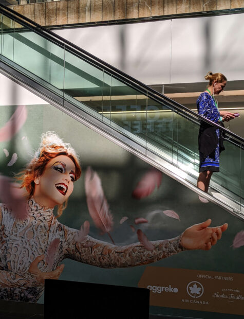 Photograph of a woman going down on an escalator. The side of the escalator has a vinyl decal of a woman with her arms open, smiling.