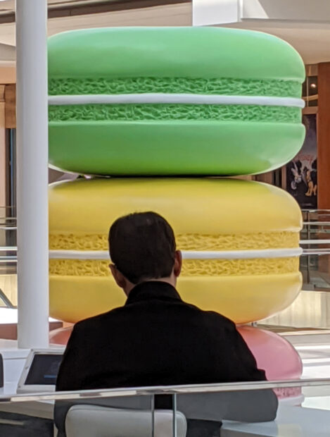 A photograph: in the foreground is a person faced away from the camera. In the background is oversized macarons in green, yellow, and pink.