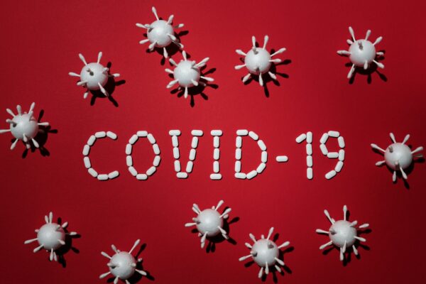 The text, "COVID-19" is spelled out with white pills on a red background. Surrounding the text are a dozen white styrofoam balls, each with several q-tips inserted into it.