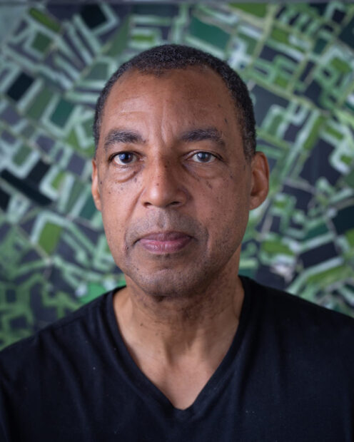A photograph of artist Rick Lowe. He looks directly at the camera and wears a black shirt. Behind him is one of his patterned paintings. 