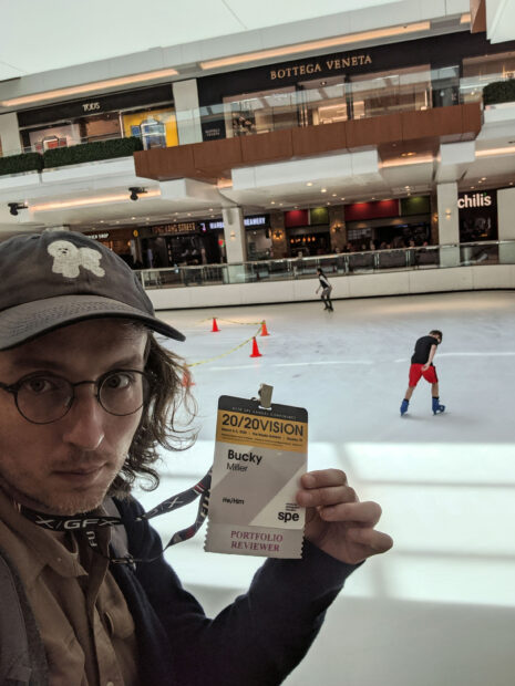 Photograph of a person wearing a hat and holding a laminated badge. In the background, there are people ice skating in a skating rink inside a mall.
