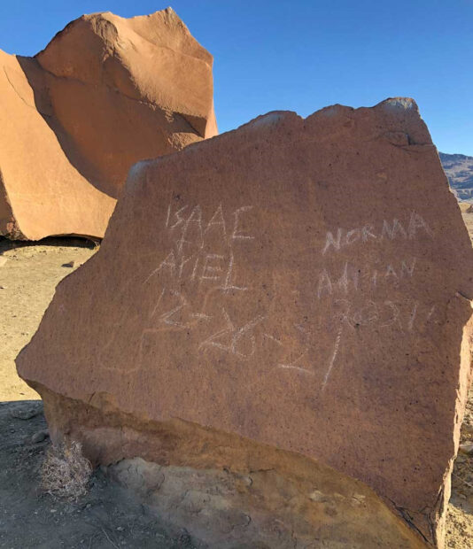 A color photograph of large red stones set against a blue sky. The stone in the foreground has a lightly etched petroglyph covered by names and a date scratched into the surface. The names are Isaac, Ariel, Norma, and Adrian. The date is 12-26-21.