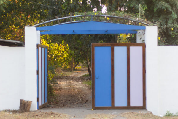 Un portón en varios tonos de azul está a medio abrir, dejando ver un sendero polvoriento al interior de una propiedad