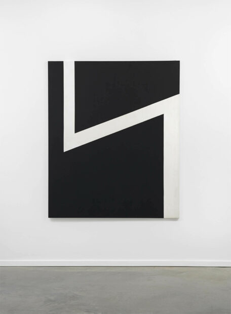 A large abstract painting on canvas by artist Carmen Herrera. The rectangular canvas is painted black with a long thin zig-zag white line that moves through the painting. The line starts at the top left, extends about halfway down, juts up and to the right edge of the canvas, and then extends straight down to the bottom right edge. The painting hangs on a white gallery wall.
