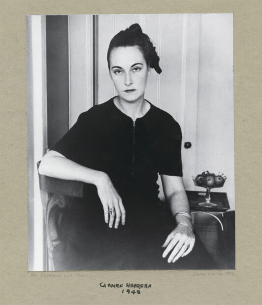 A black and white photograph of artist Carmen Herrera at age 33. She wears a short sleeve black dress and sits in a chair with one arm across her knee and one resting on the chair back. Her hair is pulled up away from her face and she looks into the camera with a serious tone.
