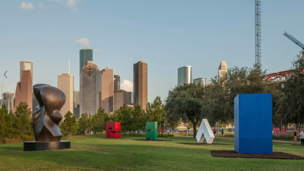 A photograph of the Buffalo Bayou Park sculpture garden with four large abstract sculptures by Carmen Herrera. The image includes a large rectangular blue sculpture, a thin white triangular sculpture, a green rectangular sculpture with triangular cutouts, and a red sculpture created from two interlocking "L" shapes. The Houston skyline is in the background.