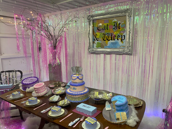 A photograph showing a close look at an installation by Sarah Ayala. The image shows a rectangular table with an array of colorful cake sculptures. The back wall of the enclosed space is decorated with pink iridescent streamers and has a framed map with hand-lettered text that reads, "Eat It & Weep."