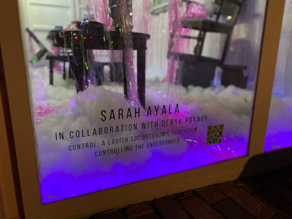 A close-up photograph of the title text on a window. The text reads, "Sarah Ayala in collaboration with Deryk Poynor: Cuntrol: A Lavish Socioeconomic Ecosystem Controlling the Underfunded." 